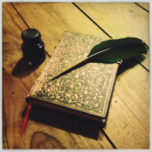 A black quill laying on an old fashioned notebook next to an ink bottle.